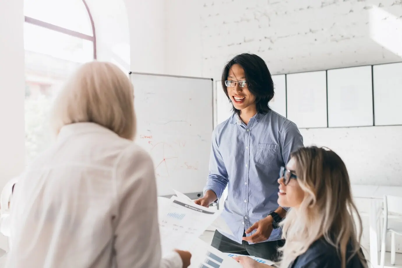 Porträt von hinten einer blonden Frau in einem modischen Hemd, die Dokumente liest, während ein asiatischer Mann ihr etwas sagt.
